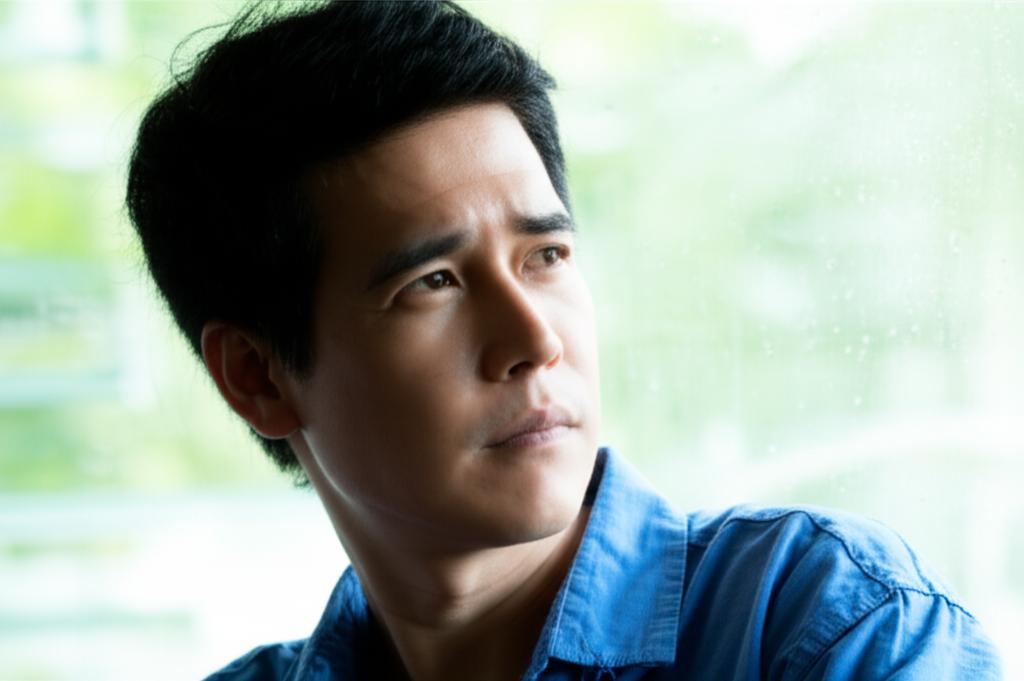 A thoughtful Thai man looking out a window, contemplating his health and future, perhaps feeling a bit tired or stressed.